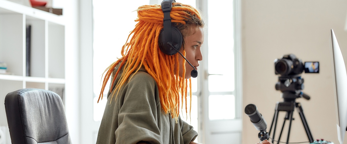 Person mit langen orangen Dreadlocks in Seitenansicht blickt auf Computermonitor, hat Kopfhörer aufgesetzt und bedient Maus und Tastatur, im Hintergrund weitere Mikrofon und Filmkamera