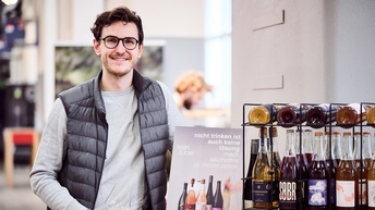 Portraitaufnahme einer Person mit einem Glas Weißwein in der rechten Hand.  Daneben ist eine hüfthohe Holztruhe , auf der ein Weinregal aus Eisen steht, in dem Weinflaschen eingeordnet sind. Daneben steht ein Aufsteller aus Papier