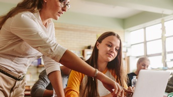 In einer Schulklasse sitzt ein Teenager an einem Tisch. Vor dem Teenager ist ein aufgeklappter Laptop, auf den sie blickt. Neben ihm steht eine erwachsene Person, die auf den Bildschirm deutet. Im Hintergrund sitzen weitere Teenager.