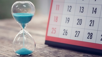 Eine Sanduhr mit blauem Sand sowie ein Kalender stehen auf einem Holztisch