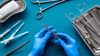 Auf einem türkischen Untergrund sind Werkzeuge für eine Operation platziert: ein Tuch, Spritzen, OP-Scheren, eine Pipette. Zwei Hände mit blauen Gummihandschuhen 
halten ein Skalpell. 