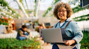 In der rechten Bildhälfte ist eine Person in einem Gewächshaus, die einen Laptop hält und direkt in die Kamera blickt. Um die Person sind viele grüne Pflanzen am Boden und auf Stangen an der Decke. Zwei weitere Personen knien im Hintergrund 