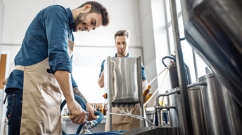 Zwei Personen mit blauem Hemd und Arbeitsschürze füllen Bier in einem Lager ab
