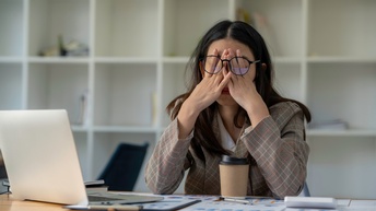 Eine Person sitzt an einem Tisch mit aufgeklappten Laptop, Unterlagen und einem Kaffeebecher. Sie hat die Hände auf ihr Gesicht gelegt