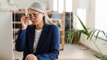 Person mit langen grauen Haaren, Brille und Businesskleidung sitzt bei einem Schreibtisch und blickt auf einen Computer-Monitor in einer hellen, modernen Büroräumlichkeit