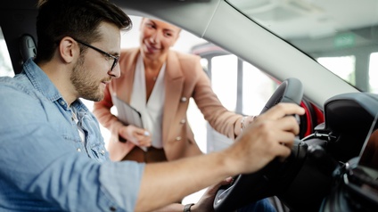Person mit Brillen sitz an Steuer eines Fahrzeugs sanft lächelnd auf Lenkrad blickend, im Hintergrund weitere Person sich über Fahrertüre leicht ins Auto neigend