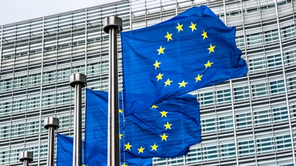 Drei EU-Flaggen im Wind wehend, blaue Stoffe mit gelben im Kreis verlaufenden Sternen an Fahnenmasten, im Hintergrund Hausfassade mit Sonnenblenden