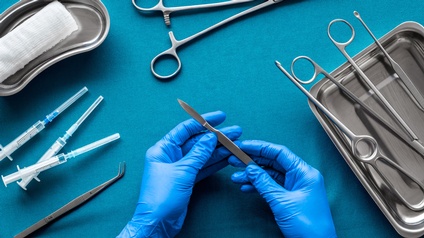 Auf einem türkischen Untergrund sind Werkzeuge für eine Operation platziert: ein Tuch, Spritzen, OP-Scheren, eine Pipette. Zwei Hände mit blauen Gummihandschuhen 
halten ein Skalpell. 
