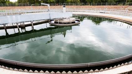 Ansicht einer Kläranlage: Großes kreisrundes Becken mit Wasser in dem sich Himmel spiegelt, im Hintergrund Zaun und Baumzeile