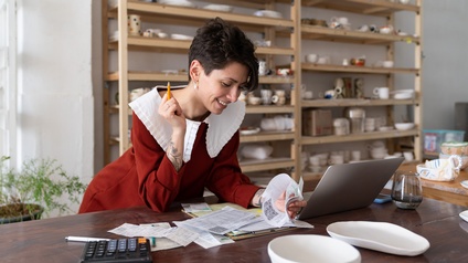 Lächelnde Person mit großem weißen Kragen lehnt über Tisch und durchblättert Zettel, hält in einer Hand Stift, ringsum am Tisch aufgeklapptes Notebook, Taschenrechner, Glas und Keramiken, im Hintergrund verschwommen Regal mit Töpferwaren