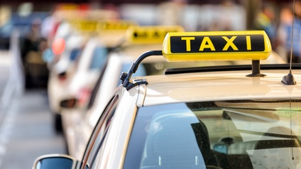 
Leuchtend gelbes Zeichen auf Dach eines beigeweißen Autos: Gelber länglicher Leuchtkörper mit schwarzen Elementen und Schriftzug Taxi , im Hintergrund verschwommen weitere parkende Taxis