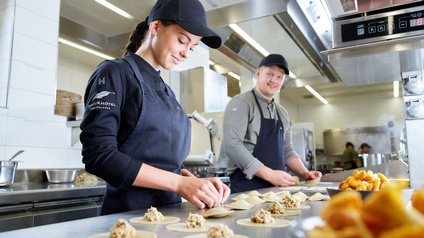 Zwei sanft lächelnde Personen mit Caps und in Schürzen stehen in großer Edelstahlküche und befüllen Teigtaschen, ringsum Küchenutensilien und Schüsseln mit Eierschwammerl