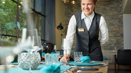 Lächelnde Person mit Zopf in grauem Gilet und weißem Hemd hält eine Hand hinter Rücken und platziert mit anderer Hand Weinglas auf gedeckter Tafel, im Hintergrund verschwommen Lichtkugeln und weitere gedeckte Tische
