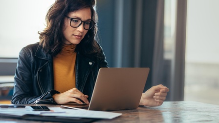 Person mit dunklen schulterlangen Haaren und Brille, schwarzer Lederjacke und gelben Pullover sitzt konzentriert vor einem Laptop an einem Tisch