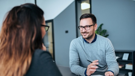 Person mit dunklen kurzen Haaren, Brille und Bart sitzt an einem Tisch und hält ein Klemmbrett sowie ein Stift in Händen während mit einer anderen Person gesprochen wird