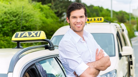 Lächelnde Person mit verschränkten Armen lehnt an Personenkraftwagen mit Taxi-Schild am Autodach, in PKW weitere Person hinter Steuer sitzend, im Hintergrund verschwommen weitere parkende Taxis