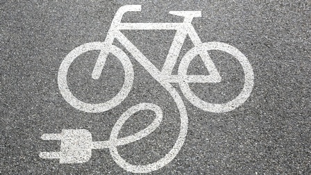 Detailansicht Asphaltstraße mit weißem aufgemalten Fahrradzeichen, Linie des Fahrrads verläuft in Zeichen für Kabel mit Stromstecker