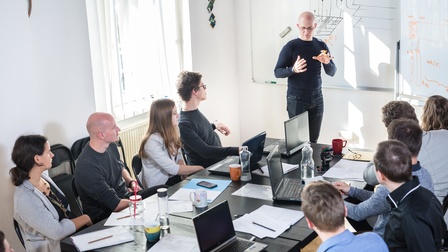 Personen nehmen an einem Meeting teil und blicken aufmerksam zur vortragenden Person auf während Laptops, Unterlagen und Trinkgefäße am Tisch stehen, im Hintergrund sind Skizzen und Notizen auf einem Whiteboard notiert