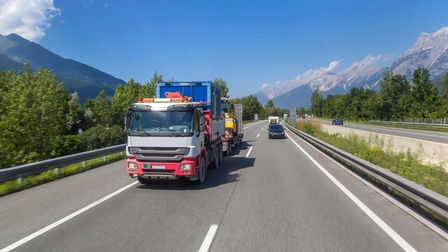 LKW und Autos fahren auf einer Autobahn, im Hintergrund sieht man abseits einen blauen Himmel sowie Bäume und Berge