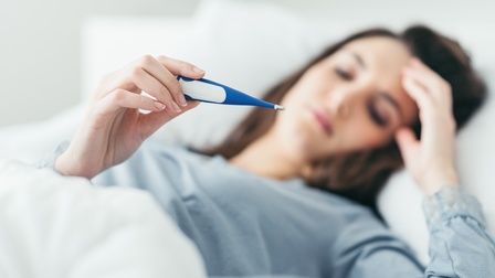Person liegt im Bett, greift sich auf den Kopf und blickt dabei auf einen Fiebermesser