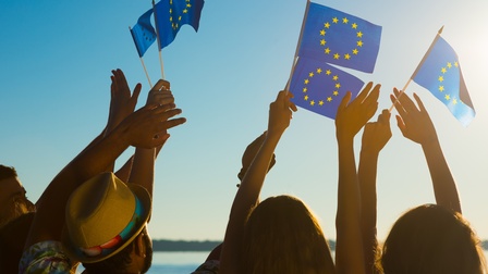 Mehrere Personen mit hochgestreckten Armen wacheln kleine EU-Flaggen in der Luft, im Hintergrund blauer Himmel