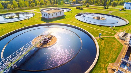 Vogelperspektive auf Kläranlage: Vier kreisrunde Wasserbecken mit Stahlkonstruktionen auf grüner Wiese, im Hintergrund grüne Baumzeilen vor Stadtlandschaft
