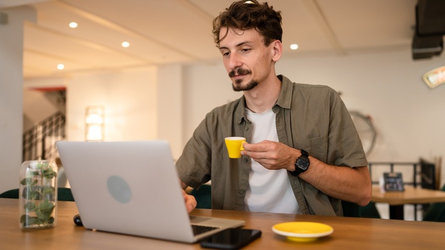 Eine Person mit dunklem Bart und kurzen, dunklen Haaren sitzt an einem Tisch. Vor ihr ist ein aufgeklappter Laptop sowie ein Smartphone und eine Untertasse. In der linken Hand hält sie eine Tasse. Die Person sitzt in einem Raum mit Spots an der Decke. 