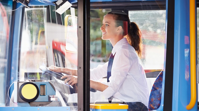 Seitliche Aufnahme einer Person in weißem Hemd und Krawatte, die hinter einem Lenkrad eines hohen Autos sitzt und geradeaus blickt. Ihre Hände sind auf dem Lenkrad. Sie hat einen dunklen Zopf