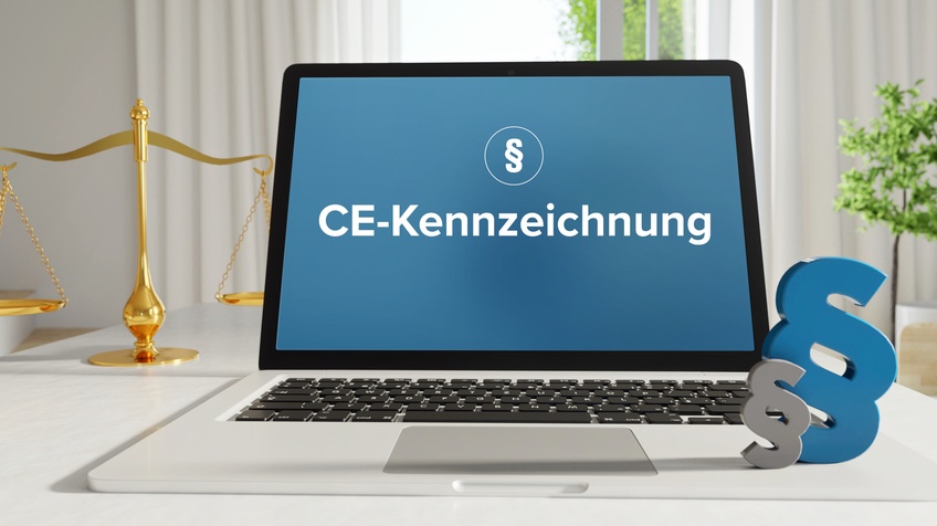Auf dem Bildschirm eines aufgeklappten Bildschirms steht in weißer Schrift CE-Kennzeichnung auf blauem Hintergrund. Auf dem Laptop sind vorne rechts zwei Paragrafenzeichen. Im Hintergrund ist eine Waage