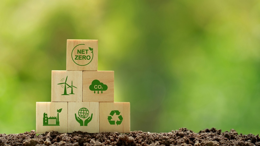 Mehrere Holzwürfel aufeinander gestapelt auf Erde mit grünen Symbolen zum Thema Klimaschutz, oberster Würfel mit Schriftzug Net Zero, Hintergrund verschwommen grün 